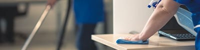 woman wiping a desk with someone else cleaning the floor in the background